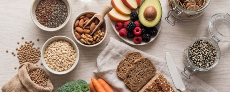 Variedad de alimentos saludables: frutas, frutos secos, panes integrales, semillas y vegetales sobre una mesa de madera clara. Ambiente fresco.