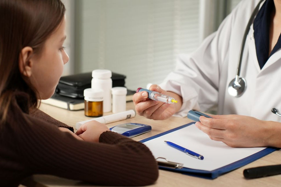 Niña observa a doctora explicando el uso de una jeringa en consultorio. Hay frascos y un bloc en la mesa. Ambiente profesional.